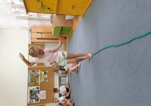 Dziewczynka ćwiczy równowagę idąc noga za nogą po rozłożonej na dywanie skakance.