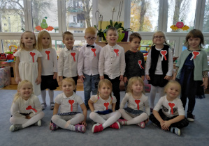 Dzieci w dwóch rzędach prezentują swoję odświętne stroje i biało-czerwone kokardy narodowe.