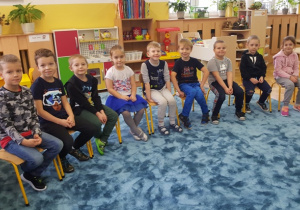 Dzieci siedzą na krzesełkach ustawionych w półkolu i czekają na rozpoczęcie spotkania.