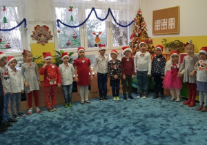 Dzieci stojąc w dużym półkolu śpiewają piosenkę dla Mikołaja.