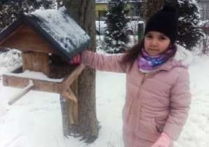 Dziewczynka dosypuje karmę dla ptaków do zawieszonego na drzewie karmnika.