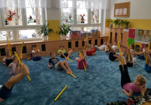 Dzieci leżąc na dywanie podnoszą stopami laskę w górę.