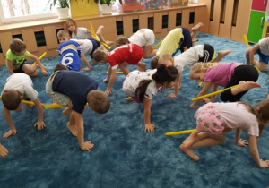 Dzieci czworakują po dywanie z laską umieszczoną pod kolanem ugiętej nogi.