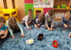 Dzieci siedzą w dużym kole. Na dywanie układają obrazy sylabowe i głoskowe wybranych słów za pomocą kolorowych krążków i koralików.
