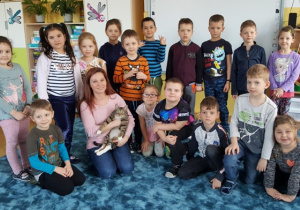 Dzieci na dywanie skupiły się wokół przybyłych gości- pani Izy i kota Simby. Robione jest pamiątkowe zdjęcie.