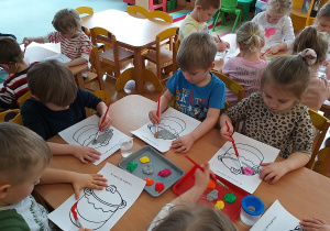 Dzieci siedzą przy stolikach i zamalowują farbami garnek.