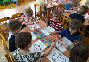 Dzieci siedzą przy stolikach i zamalowują farbami garnek.