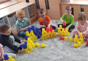 Dzieci siedzą na dywanie i układają własne wieże z kolorowych kubeczków.