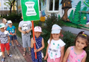 Dzieci idą zamieścić przed przedszkolem tabliczkę z napisem "Prosimy nie deptać trawnika"".