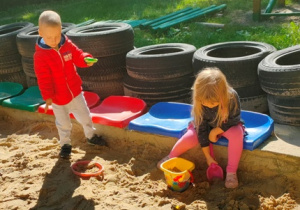 Dwoje dzieci buduje formy z piasku w dużej piaskownicy.