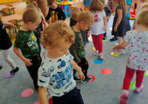 Dzieci przy dźwiękach muzyki maszerują po kropkach rozłożonych na dywanie.