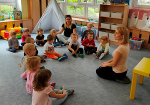 Dzieci siedząc na dywanie wykonują ćwiczenia oddechowe.