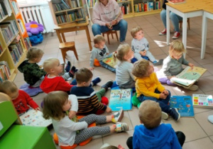 Dzieci siedzą na kolorowych poduszkach ułożonych na podłodze i oglądają książeczki.