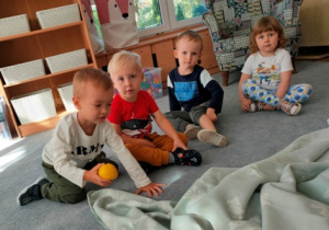 Dzieci siedzą na dywanie- pod kocem po kolei szukają zabawkowych owoców.
