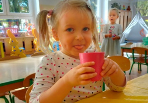 Dziewczynka siedząc przy stole pije sok.