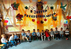 Dzieci siedzą na krzesełkach ustawionych w półkolu na dywanie- czekają na rozpoczęcie uroczystości.