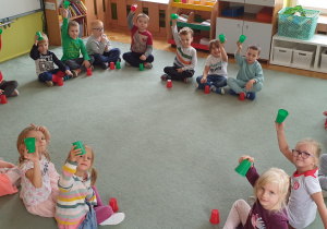 Dzieci siedzą w kole na dywanie i biorą udział w zabawie z kolorowymi kubeczkami.