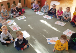 Dzieci siedzą na dywanie w kole i prezentują swoje prace plastyczne, które wykonał wspólnie w parach.