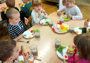 Dzieci siedząc przy stoliku jedzą kanapki przygotowane przez siebie.