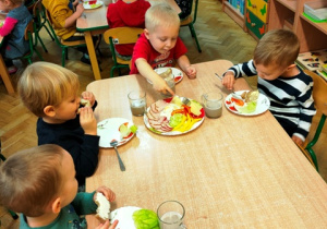 Chłopcy siedząc przy stoliku jedzą z apetytem kanapki, jedno dziecko dobiera z talerza żółty ser na swoją kanapkę.