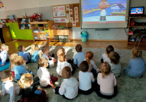 Dzieci siedząc na dywanie oglądają bajkę edukacyjną "Symbole Narodowe" wyświetloną na tablicy multimedialnej.