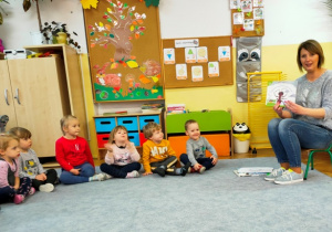 Dzieci siedzą w półkolu na dywanie. Ciocia Ania prezentuje dzieciom książkę o mrówce Mądralinie.