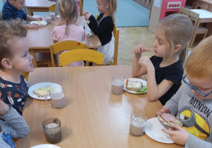 Dzieci siedzą przy stolikach i jedzą śniadanie, które same przygotowały.