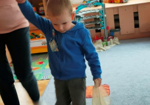 Chłopiec pokonuje tor przeszkód- maszeruje po chusteczkach ułożonych na dywanie.