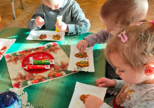 Dzieci siedzą przy stoliku i dekorują pierniczki pisakami cukrowymi i posypką cukrową.